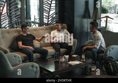 Three asian young men sitting in coffee shop, drinking coffee, talking and laughing. Design in coffee shop, restaurant, group of young people Stock Photo