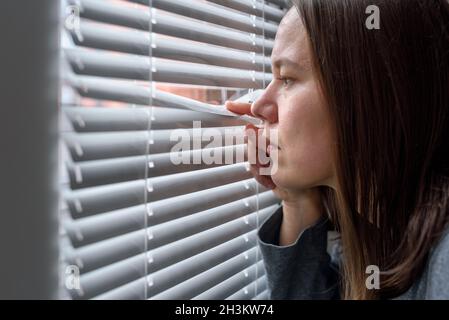 Anxious woman looking to window through blinds and waiting Stock Photo