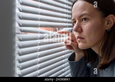 Anxious woman looking to window through blinds and waiting Stock Photo