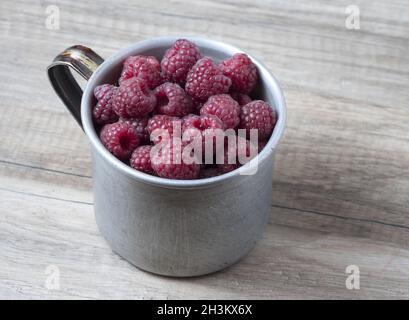 Ripe sweet raspberries in old rustic metal cup on wooden background. Stock Photo