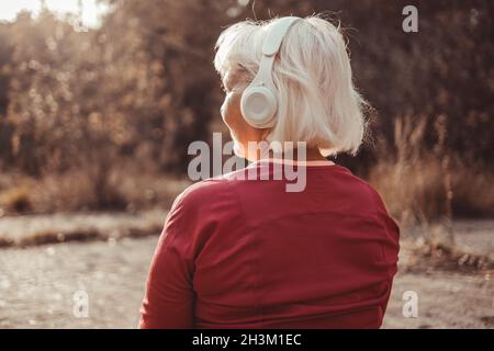 Back view of caucasian woman listening music with headphones on sunny day while walking in city park Stock Photo