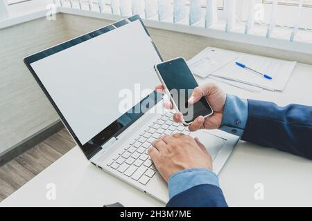 Male hands using smartphone mockup with blank screen at office desk with an open laptop mockup and tablet computer. For responsive design presentation Stock Photo