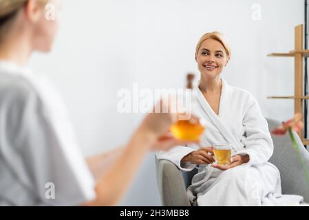 happy woman in bathrobe holding cup of tea while looking at blurred spa specialist with jug of massage oil Stock Photo