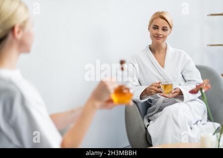 joyful woman in bathrobe holding cup of tea while looking at blurred spa specialist with massage oil in jug Stock Photo