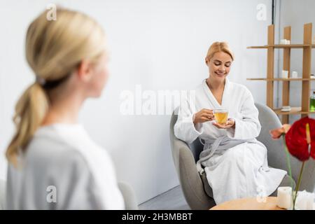 cheerful woman in bathrobe holding cup of tea while sitting near blurred spa specialist Stock Photo