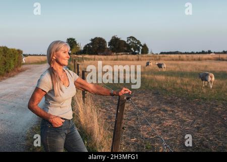 Smiling senior female farmer standing by fence while looking at field Stock Photo