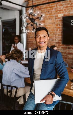 Portrait of businessman standing with laptop while colleagues discussing in background Stock Photo