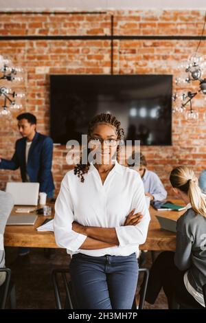 Portrait of confident female professional standing with arms crossed while colleagues discussing in background Stock Photo