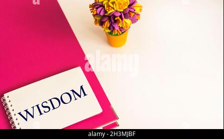 The word Wisdom is written on a notebook that lies on a pink folder, next to a bright bouquet of dried flowers on a white background Stock Photo
