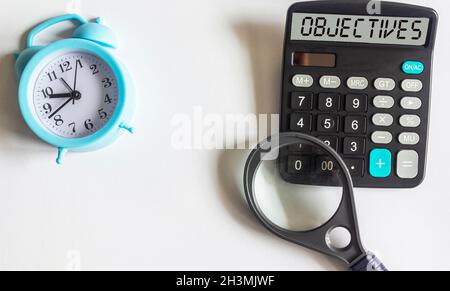 the text OBJECTIVE is written on the display of the calculator, next to a magnifying glass and an alarm clock on a white background. Place to write Stock Photo