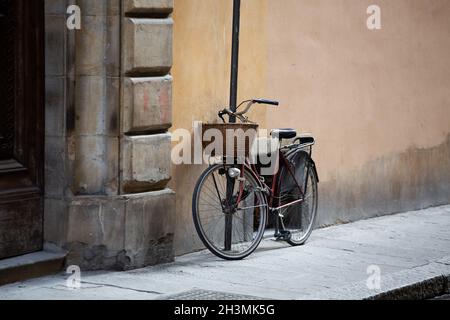Bicycle standing near wall of ancient building. Female bike with wicker basket in front is fastened with chain to post on street Stock Photo