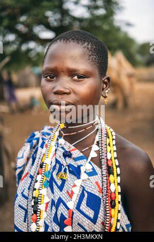 TOPOSA TRIBE, SOUTH SUDAN - MARCH 12, 2020: Teen girl in ornamental garment and with colorful accessories looking at camera on b Stock Photo