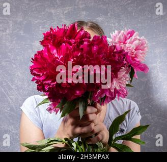 Large blurred bouquet of pink peonies covering face young woman on grey. Focus on flowers. Trendy style without face. Impersonal image. Faceless trend. Stock Photo