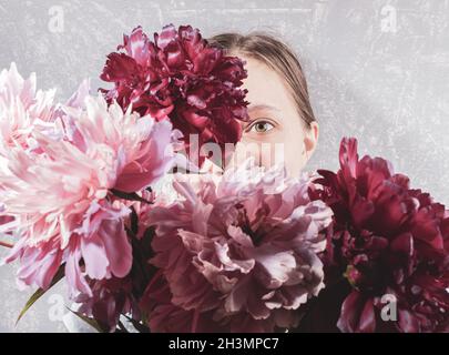 Large blurred bouquet of pink peonies covering face young woman on grey background. Focus are on one her eye. Trendy style without face. Creative retr Stock Photo