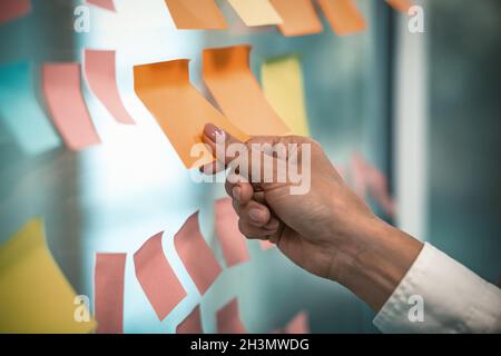 Female hand glues empty sticker on office window. Many colorful note paper stickers with space for text pasted on glass wall. Ti Stock Photo