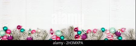 Christmas border banner with pink, purple and teal decorations and tree branches. Overhead view on a white wood background. Stock Photo