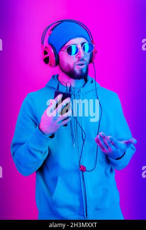 Funky young bearded hipster man in glasses and headphones dances and listens pleasurable music in his smartphone. love live. Lei Stock Photo