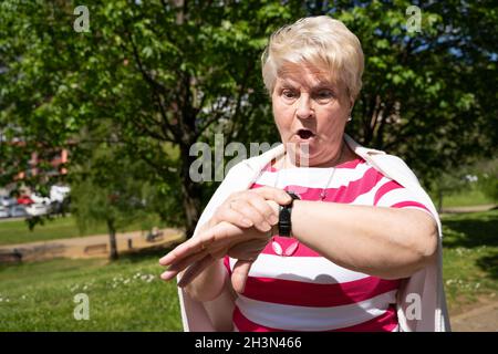 Elder woman looking at watch with surprise look at the park. Senior blonde lady realizing she is late on sunny day outdoors Stock Photo