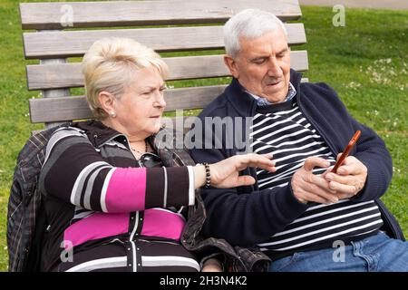 Senior couple sitting on bench struggling with mobile phone. Elder husband and wife using cellphone at park Stock Photo