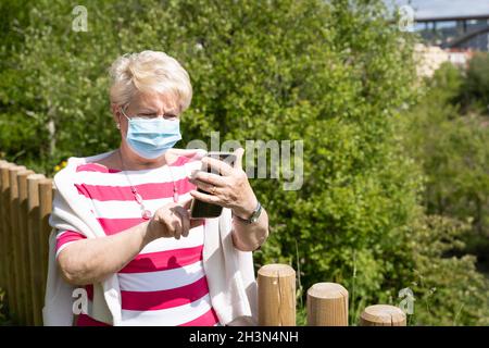 Senior woman with face mask touching cellphone screen at park on sunny day. Elder lady standing using mobile phone on natural landscape Stock Photo