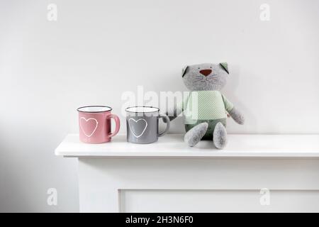 A pink and gray mug with a painted white heart, two funny toy rag cats are positioned on the fireplace. Home decoration for Valentine's Day. Scandinav Stock Photo