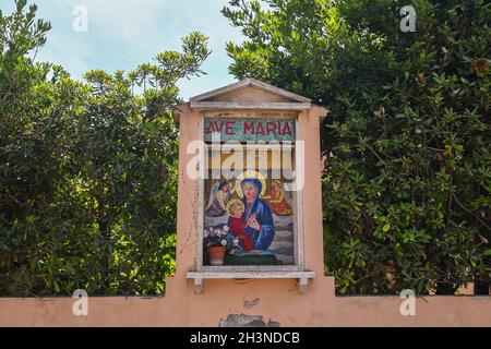 Colorful mosaic shrine depicting Saint Mary with Child Jesus on the enclosing wall of a garden with leafy plants in summer, San Vincenzo, Livorno Stock Photo