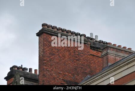 A large row of old fashioned traditional clay chimney pots on a red brick support against a grey sky from when buildings where b Stock Photo