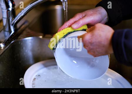 Senior man hands wash plate dishes in kitchen sink under running water at home. Authentic senior lifestyle after retirement and household work. Stock Photo