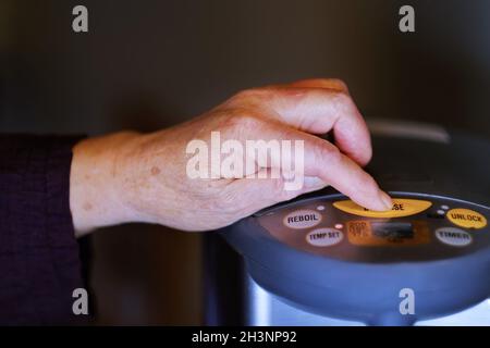 Close up of senior woman operating electric Hot Water Boiler to dispense hot water at home. Authentic senior retirement modern lifestyle background. Stock Photo