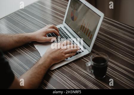 Close-up of a man who is typing on a laptop and working with a finance charts on a messy wood office table. Stock Photo