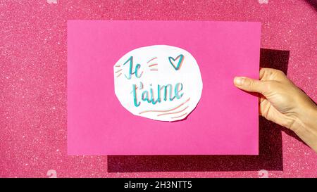 handwritten sign on white paper pasted on a pink cardboard held by one hand with the words je t' aime Stock Photo