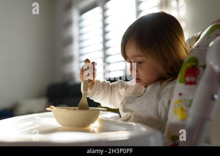 Small girl eats brakefast by herself with a spoon. Stock Photo