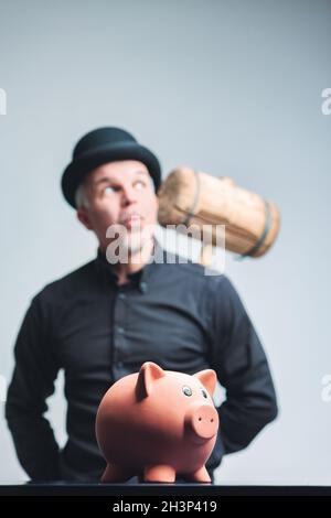 Senior man in bowler hat contemplating smashing his piggy bank holding a large mallet behind his back with focus to the pig in the foreground Stock Photo