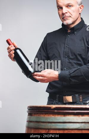 Senior man or sommelier standing holding a bottle of red wine behind an old oak cask or barrel in a cellar in a viticulture concept Stock Photo
