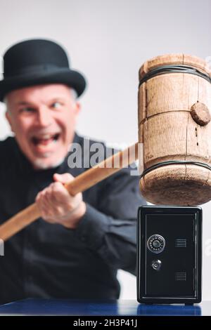 Gleeful senior man in bowler hat trying to smash a small metal safe or piggy bank with a large wooden mallet in a conceptual image with focus to the s Stock Photo