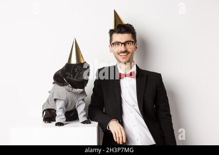 Image of handsome young man celebrating birthday with cute black pug in party costume and cone on head, standing over white back Stock Photo