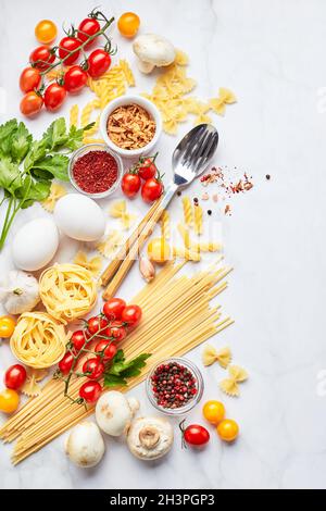 Food background with different kinds of pasta, tomatoes, herbs, mushrooms, eggs, seasonings scattered on light marble background Stock Photo