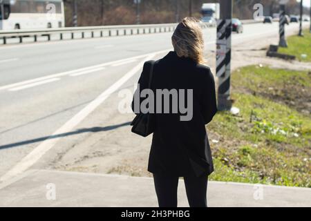 A girl in strict black clothes with a bag is waiting for the bus at the bus stop, rear view. Sunny day out of town. Late autumn. Traffic. City Lifesty Stock Photo