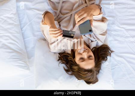 Woman lying in bed makes an order through a mobile application. She dials the card number for online purchases. Online shopping concept. View from the Stock Photo