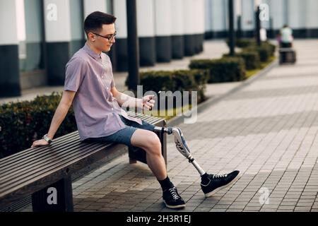 Disabled young man with foot prosthesis sitting on bench on city street and holds mobile phone outdoor. Stock Photo