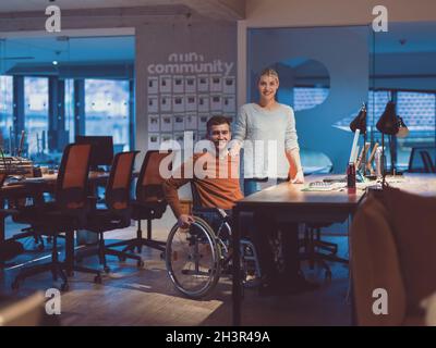 Businessman in a wheelchair in modern coworking office space working late night in office. Colleagues in background. Disability Stock Photo