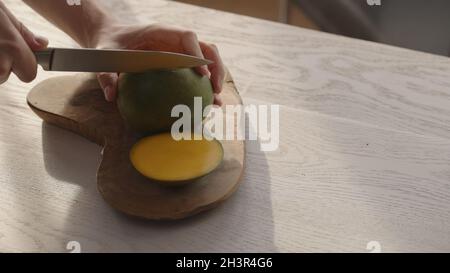 Man cutting ripe mango on olive board on white oak table, wide photo Stock Photo