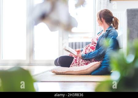 Preparing for pregnancy: Pregnant Caucasian mother is sitting on the floor, reading a book for about babies Stock Photo