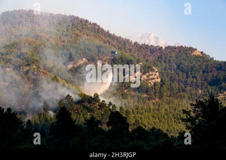 Wildfire helicopter drops water on a fire in a steep, rocky terrain with a bucket of water. The mountains are covered with dense pine forests. Stock Photo