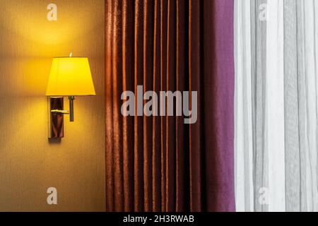 A combination of a modern wall sconce, a classical patterned curtain and a tulle curtain. The light is on. Sunlight is coming from outside. Stock Photo