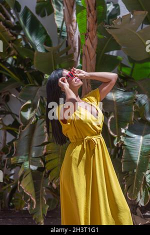 Vertical image of a Latina woman in a yellow dress wearing sunglasses and holding them with both hands looking up against a background of green plants Stock Photo