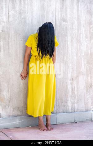 Vertical image of an adult woman with long black hair barefoot and standing leaning on a wall with her head tilted forward and her hair covering her f Stock Photo
