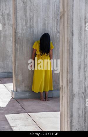 Vertical image of an adult woman with a yellow dress with long black hair covering her face barefoot leaning on a wall or column at daytime. Stock Photo