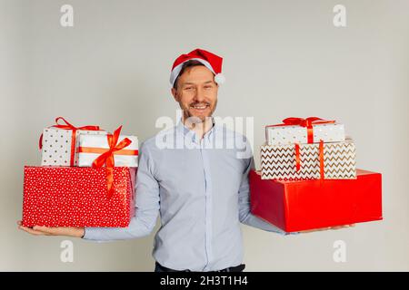 Handsome surprised businessman man in red Santa hat holding many gift boxes, He has happy smile on face, ready for holidays against grey background. Stock Photo