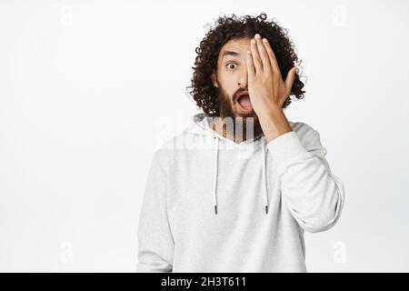 Portrait of guy covers half of face with palm, looks surprised and amazed at camera with one eye, white background Stock Photo